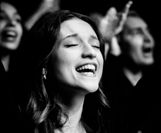 Hier ist eine singende Frau zusehen. Im Hintergund sind noch andere Menschen zu sehen, die auch singen.