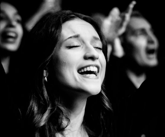 Hier ist eine singende Frau zusehen. Im Hintergund sind noch andere Menschen zu sehen, die auch singen.