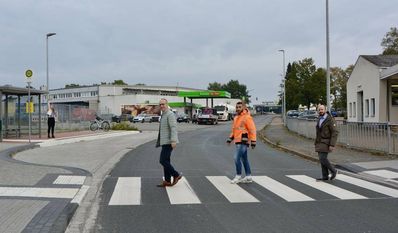 Während eine Person an der Haltestelle in der Industriestraße wartet, spazieren Baudezernent Frank Quell (v. li.), B.Eng. Wael Youssef und Fachbereichsleiter Tobias Bersch in „Beatles-Manier“ (siehe Albumcover „Abbey Road“) über den neuen Zebrastreifen zur Umgebauten Bushaltestelle.