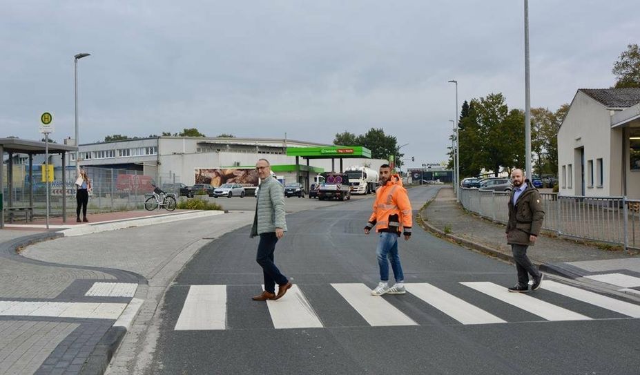 Während eine Person an der Haltestelle in der Industriestraße wartet, spazieren Baudezernent Frank Quell (v. li.), B.Eng. Wael Youssef und Fachbereichsleiter Tobias Bersch in „Beatles-Manier“ (siehe Albumcover „Abbey Road“) über den neuen Zebrastreifen zur Umgebauten Bushaltestelle.