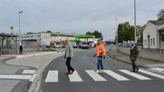 Während eine Person an der Haltestelle in der Industriestraße wartet, spazieren Baudezernent Frank Quell (v. li.), B.Eng. Wael Youssef und Fachbereichsleiter Tobias Bersch in „Beatles-Manier“ (siehe Albumcover „Abbey Road“) über den neuen Zebrastreifen zur Umgebauten Bushaltestelle.