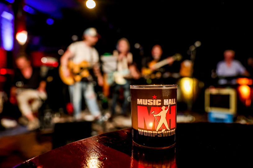 Foto der Music Hall Session, vorne eine beleuchtetes Glas, hinten leicht verschwommen Musiker