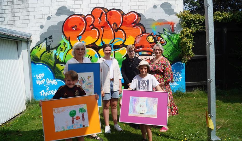 Die stolzen Preisträger:innen vor dem ebenfalls während des Kultursommers entstandenen Grafitto, v. l. n. r.: hinten: Wiebke Müller mit dem Bild von Eva Henjes, Leitung kidsArt, Luna Kolakowski (15), Aiko Drescher (23), Beate Schröder, Leiterin der Schreibwerkstatt, vorne Theo Busch (6) und Frederike Hahn (8). Nicht im Bild: Peter Sperling und Eva Henjes