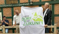 Ralf Gebken, Leiter der Jury, übergibt an der Meyenburger Wassermühle Ortsbürgermeister Dominik Schmengler einen großen Banner: „Unser Dorf hat Zukunft“.
