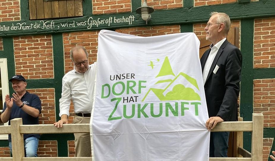 Ralf Gebken, Leiter der Jury, übergibt an der Meyenburger Wassermühle Ortsbürgermeister Dominik Schmengler einen großen Banner: „Unser Dorf hat Zukunft“.