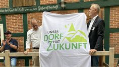 Ralf Gebken, Leiter der Jury, übergibt an der Meyenburger Wassermühle Ortsbürgermeister Dominik Schmengler einen großen Banner: „Unser Dorf hat Zukunft“.